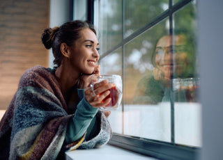 Young smiling woman drinking tea and looking through a window at home.