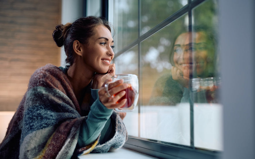 Young smiling woman drinking tea and looking through a window at home.