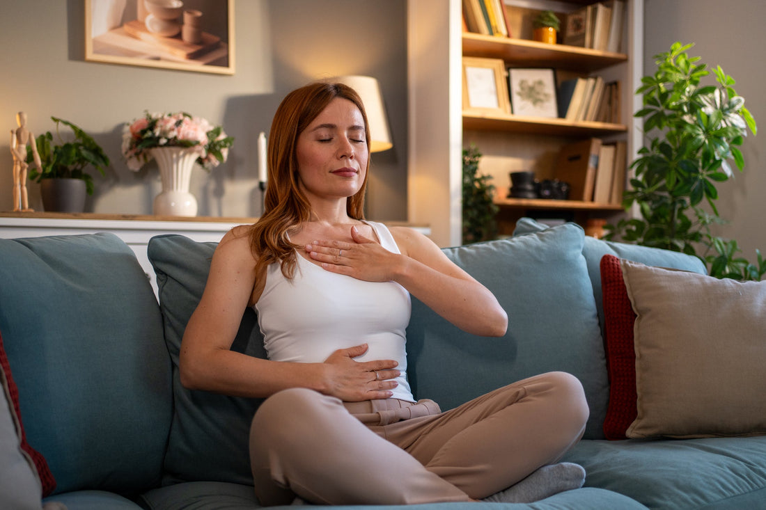 Young adult woman sitting in a lotus position on a comfortable couch, practicing deep breathing with her eyes closed.