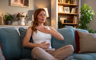Young adult woman sitting in a lotus position on a comfortable couch, practicing deep breathing with her eyes closed.