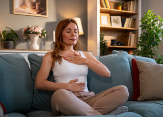 Young adult woman sitting in a lotus position on a comfortable couch, practicing deep breathing with her eyes closed.