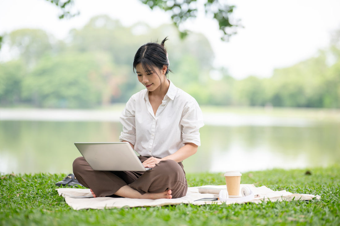 Woman in a white shirt using a laptop while sitting on a blanket on the grass.