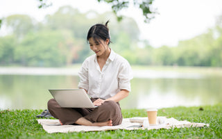 Woman in a white shirt using a laptop while sitting on a blanket on the grass.