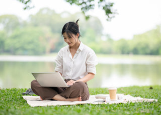 Woman in a white shirt using a laptop while sitting on a blanket on the grass.