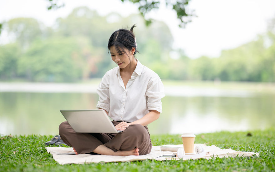 Woman in a white shirt using a laptop while sitting on a blanket on the grass.