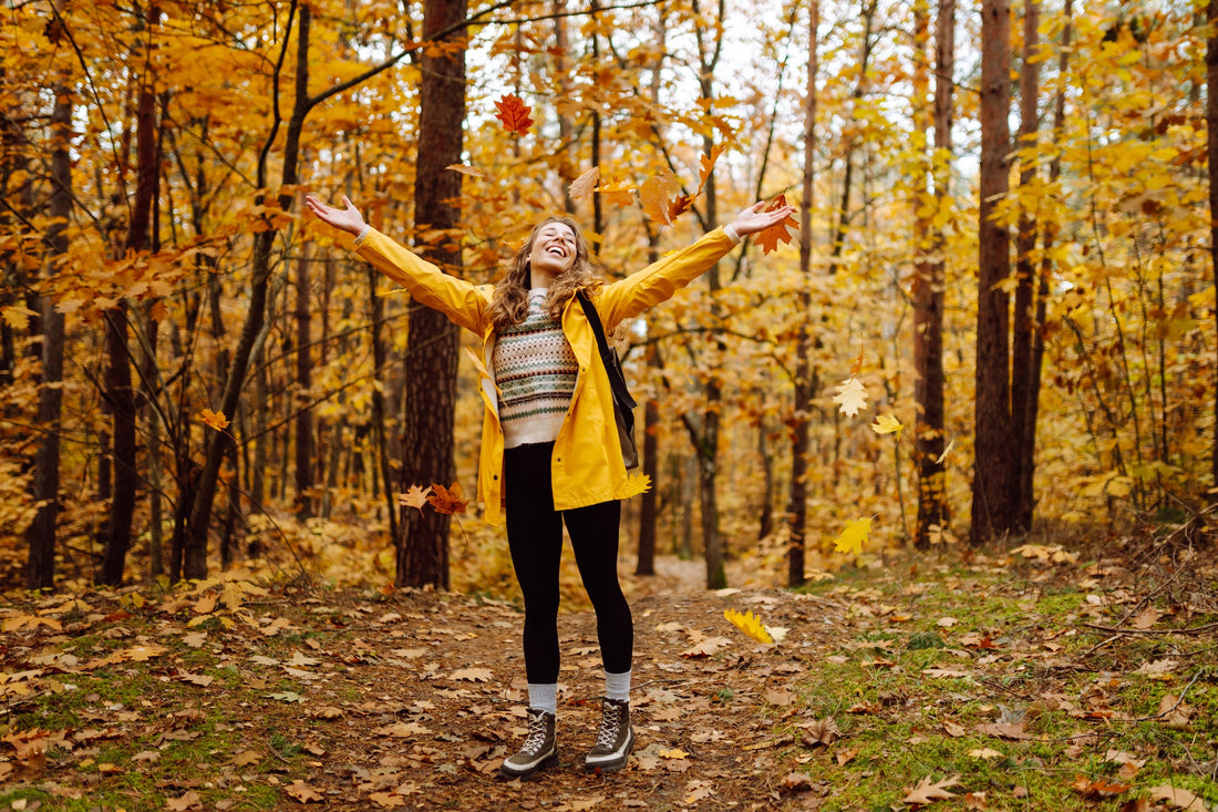 A young woman in a yellow jacket throwing leaves up above her head. 