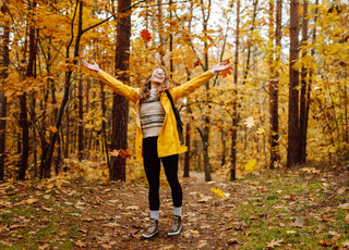 A young woman in a yellow jacket throwing leaves up above her head. 