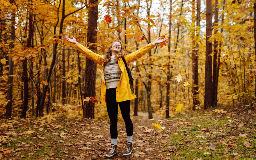 A young woman in a yellow jacket throwing leaves up above her head. 