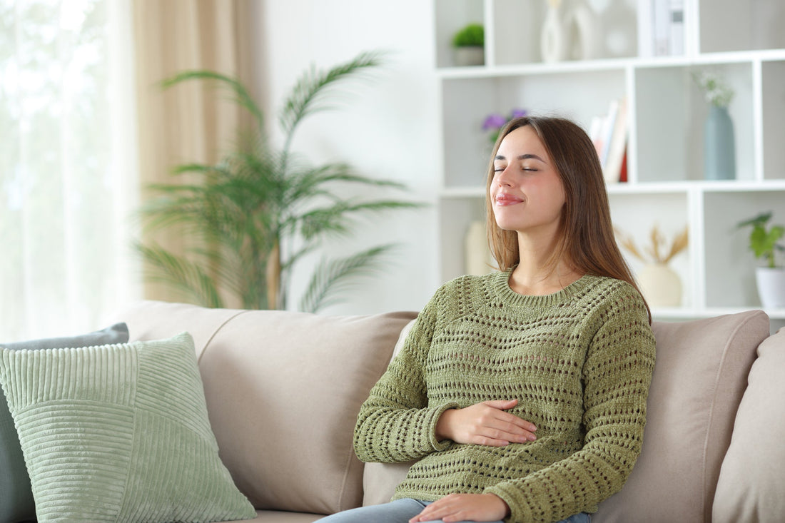 Content woman wearing a green sweater while sitting on the couch at home with one hand placed on her stomach.