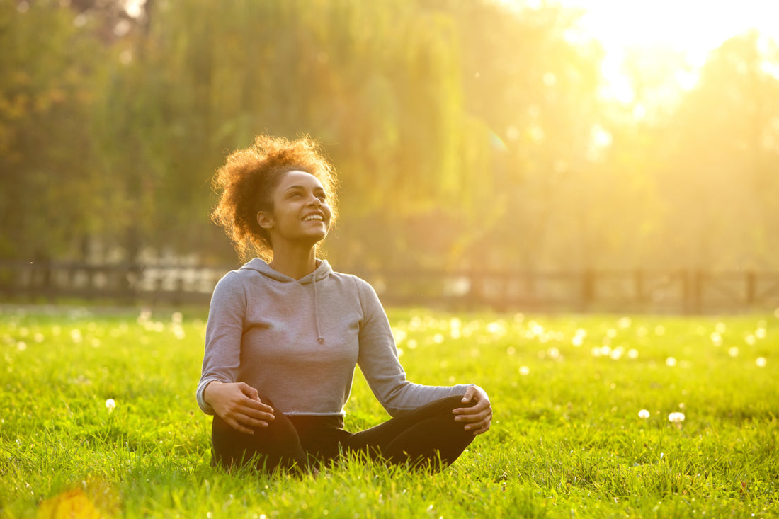 Woman in black leggings and grey sweater sitting cross-legged in the grass smiling up at the setting sun.