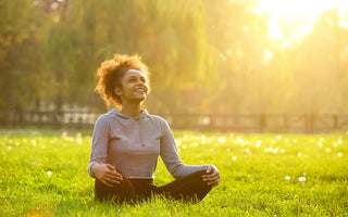 Woman in black leggings and grey sweater sitting cross-legged in the grass smiling up at the setting sun.