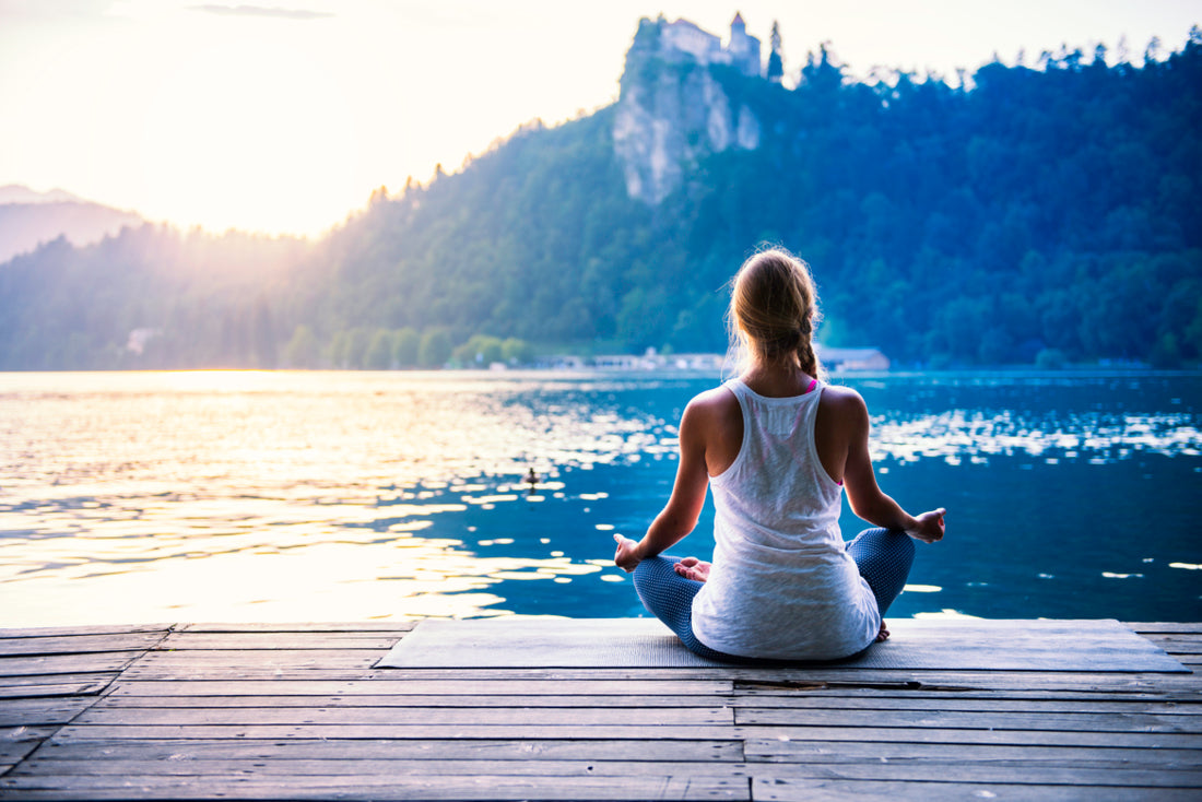 Woman in gray leggings and a white tank top is sitting cross-legged at the end of a dock on a lake as if she is meditating.