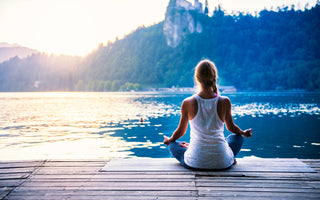 Woman in gray leggings and a white tank top is sitting cross-legged at the end of a dock on a lake as if she is meditating.