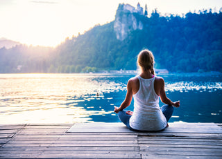 Woman in gray leggings and a white tank top is sitting cross-legged at the end of a dock on a lake as if she is meditating.