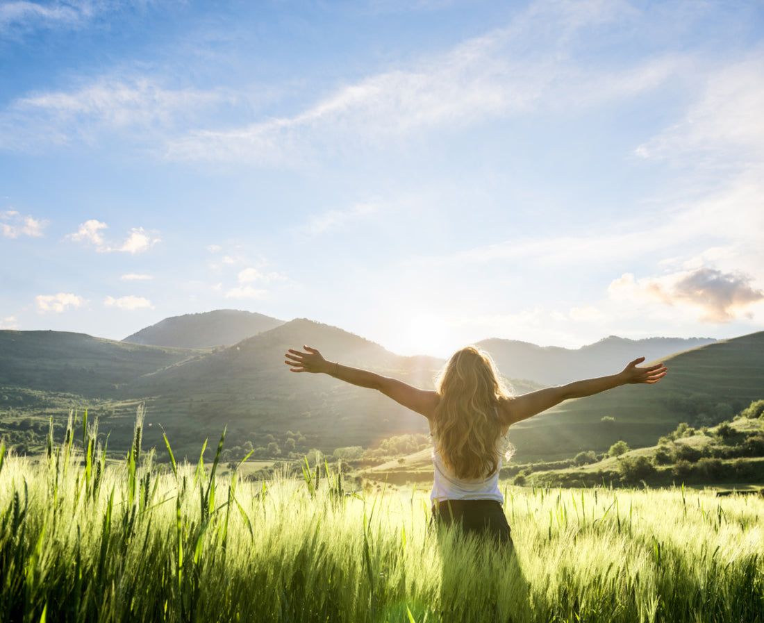 Woman standing in a field of tall grass with her arms spread out wide while she takes in the view of the mountains in front of her.