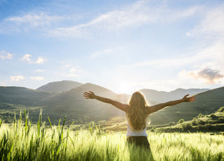 Woman standing in a field of tall grass with her arms spread out wide while she takes in the view of the mountains in front of her.