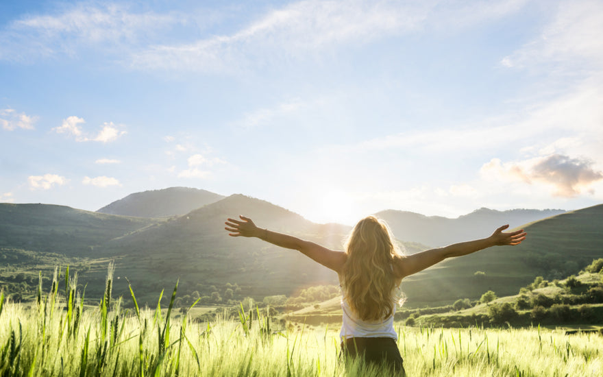 Woman standing in a field of tall grass with her arms spread out wide while she takes in the view of the mountains in front of her.