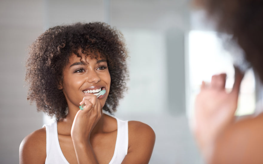 Woman in a white tank top looking at herself in the mirror while she brushes her teeth.