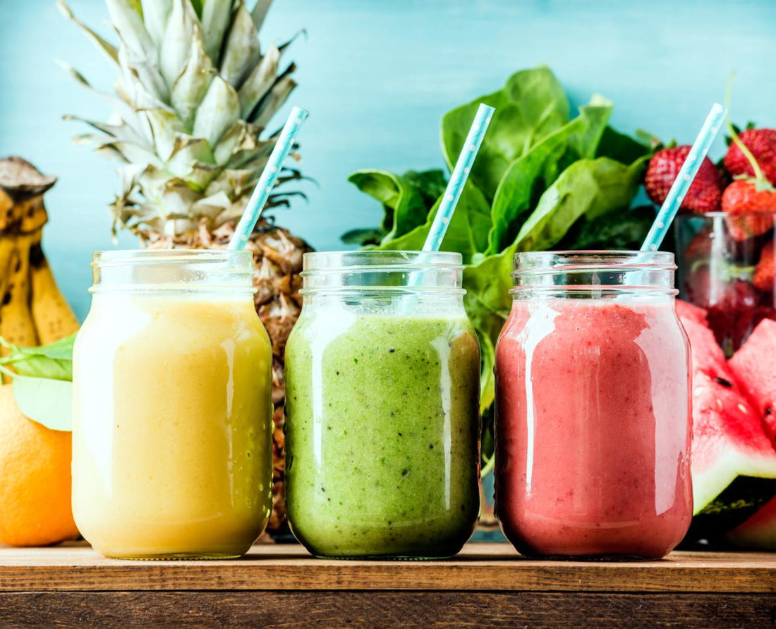 Three mason jars full of colorful smoothies sitting on a wooden table in front of fresh smoothie ingredients like pineapple and spinach.