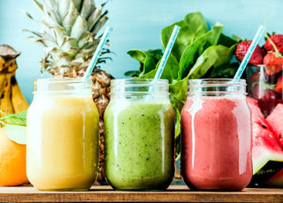 Three mason jars full of colorful smoothies sitting on a wooden table in front of fresh smoothie ingredients like pineapple and spinach.