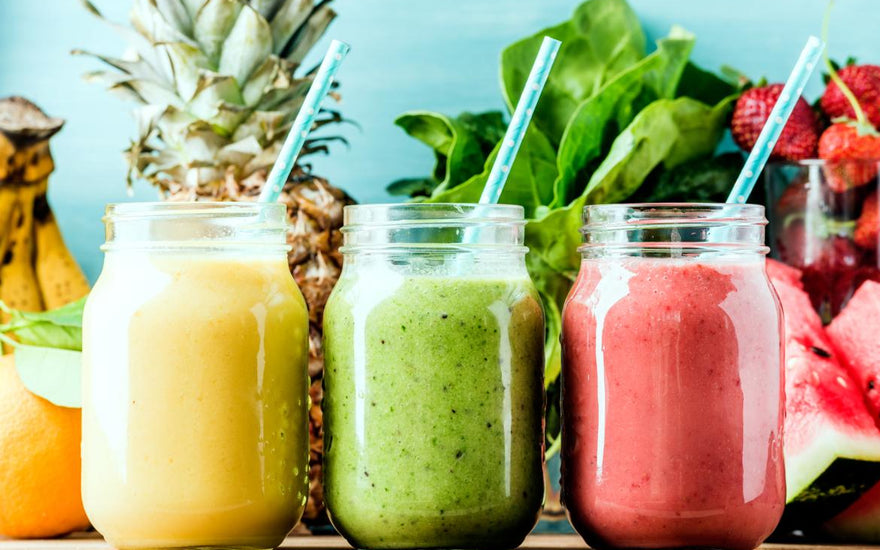 Three mason jars full of colorful smoothies sitting on a wooden table in front of fresh smoothie ingredients like pineapple and spinach.