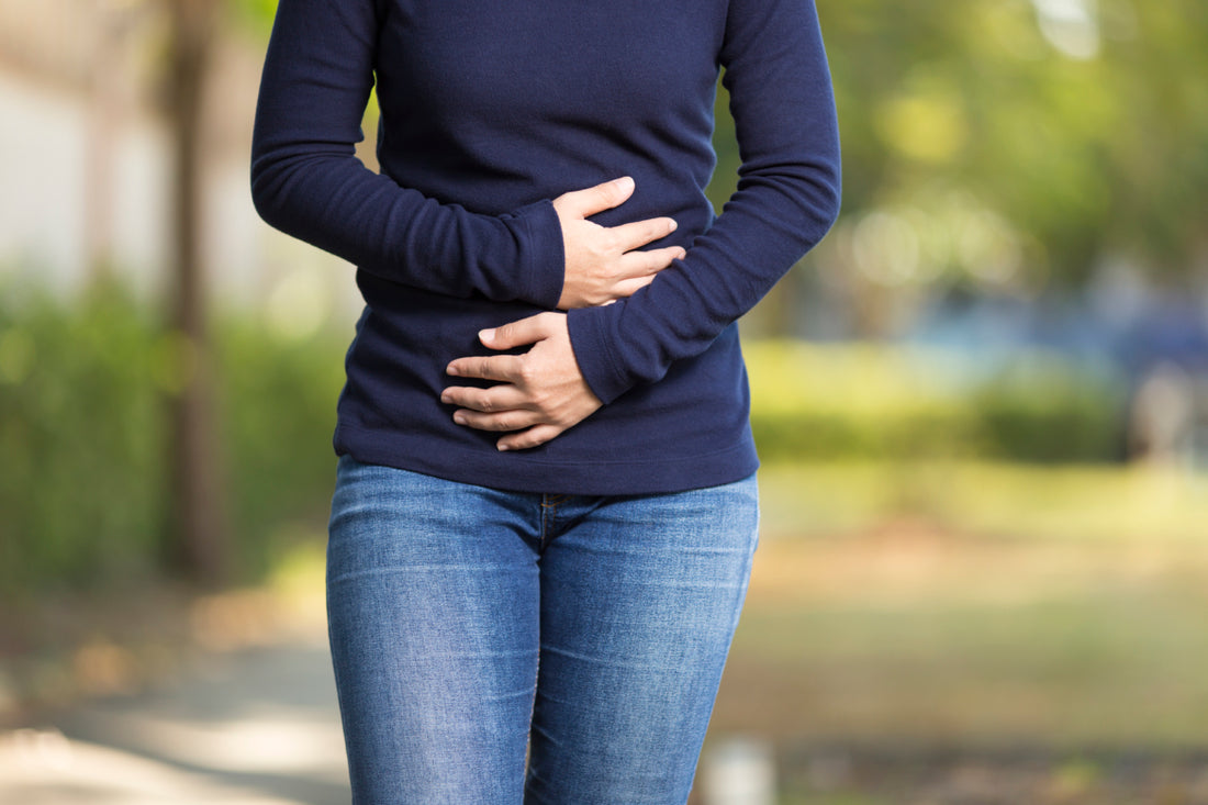 Woman in a blue long sleeve shirt and jeans standing outside holding her hands over her stomach as if she is uncomfortable.