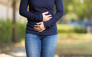 Woman in a blue long sleeve shirt and jeans standing outside holding her hands over her stomach as if she is uncomfortable.