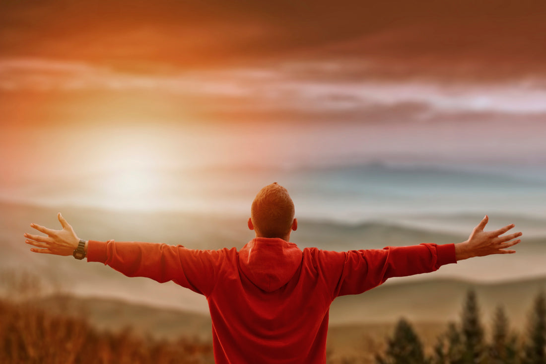 Man in a red sweater holding his arms to his sides while looking out over the sunset and wilderness landscape.