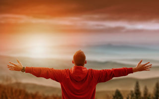 Man in a red sweater holding his arms to his sides while looking out over the sunset and wilderness landscape.