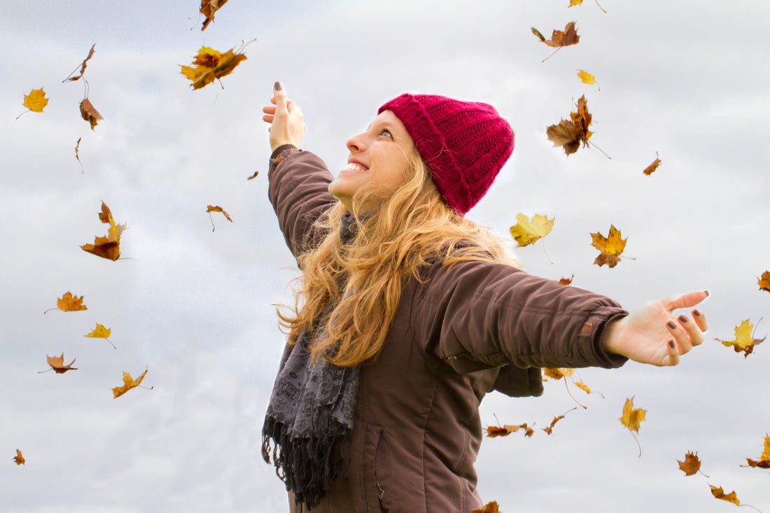 Woman in a brown jacket, black scarf, and red beanie standing with her arms spread out as autumn leaves fall around her.