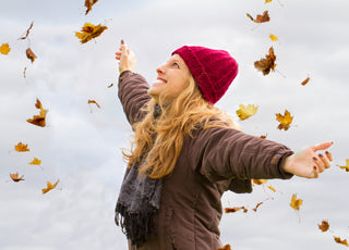 Woman in a brown jacket, black scarf, and red beanie standing with her arms spread out as autumn leaves fall around her.