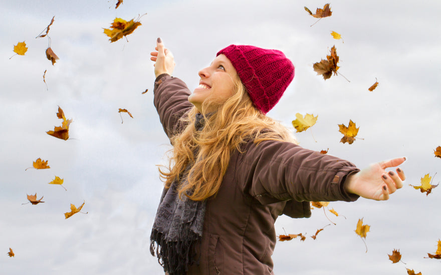 Woman in a brown jacket, black scarf, and red beanie standing with her arms spread out as autumn leaves fall around her.