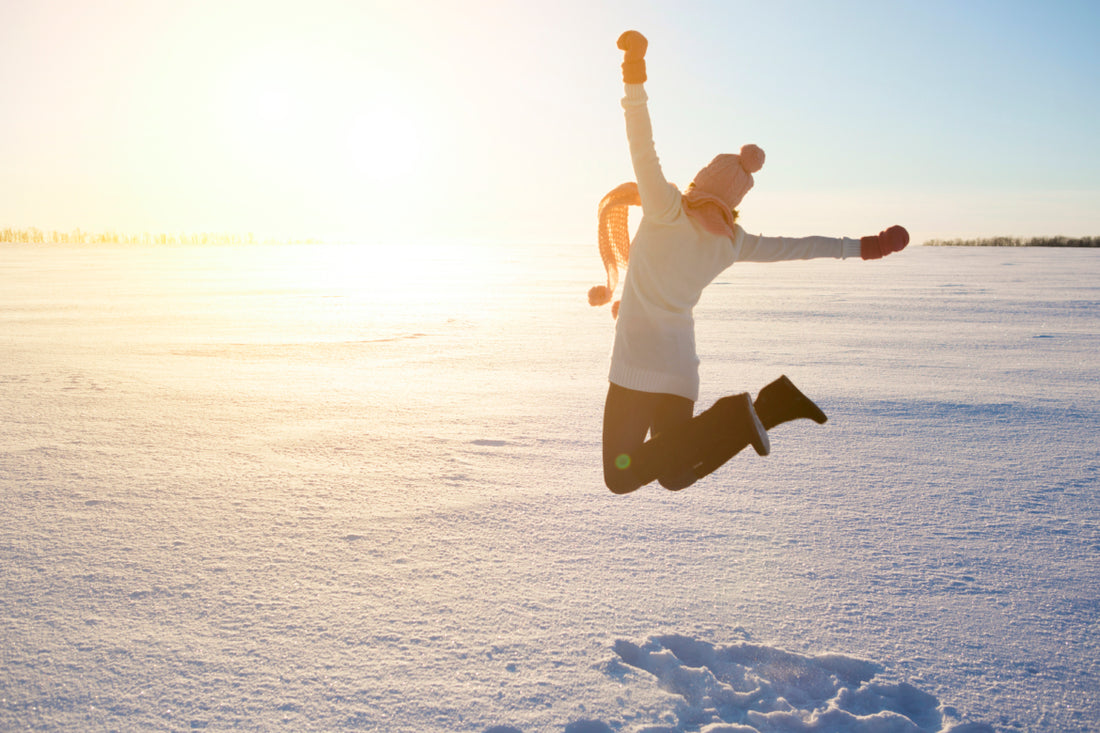 Female dressed in winter gear jumping into the air in front of a snowy sunset.