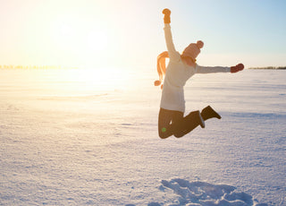 Female dressed in winter gear jumping into the air in front of a snowy sunset.