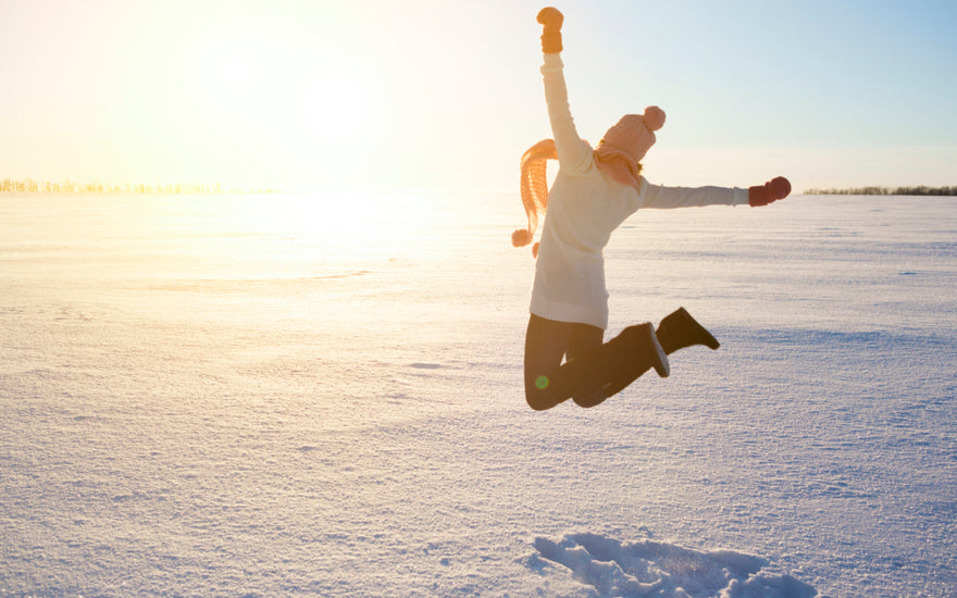 Female dressed in winter gear jumping into the air in front of a snowy sunset.