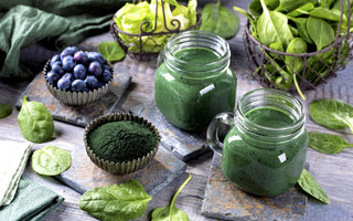 Two green smoothies sitting on a wooden table surrounded by smoothie ingredients like spinach, blueberries, and green algae powder.