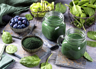 Two green smoothies sitting on a wooden table surrounded by smoothie ingredients like spinach, blueberries, and green algae powder.