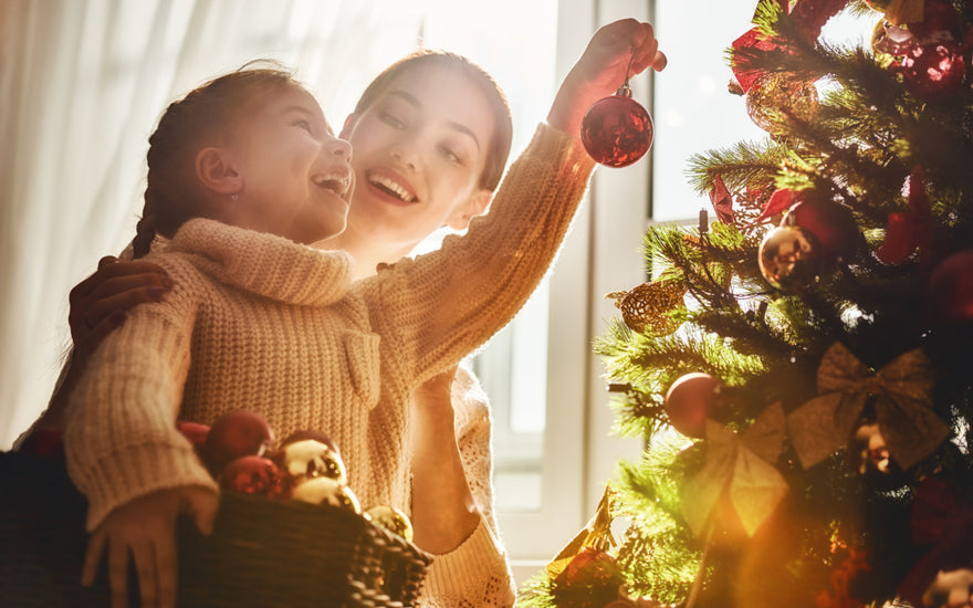 Mother and daughter in matching sweaters smiling while hanging ornaments on the Christmas tree together.