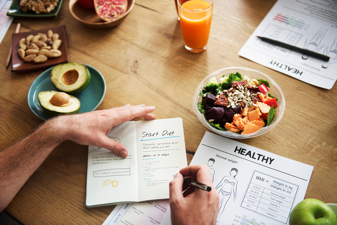 Top down view of a wooden table top with an array of healthy foods around as someone writes notes in their wellness journal. 