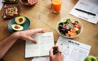 Top down view of a wooden table top with an array of healthy foods around as someone writes notes in their wellness journal. 