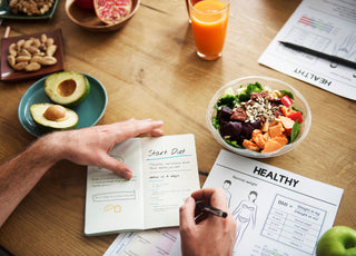 Top down view of a wooden table top with an array of healthy foods around as someone writes notes in their wellness journal. 