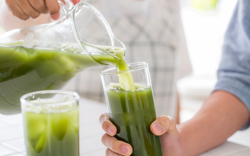 One person holding a pitcher full of homemade green juice pouring some of the contents into a glass that is held by someone else. 