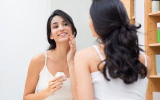 Woman in a white tank top looking at herself in the mirror as she applies moisturizer to her face.