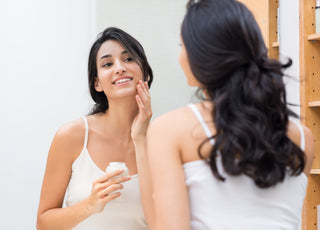 Woman in a white tank top looking at herself in the mirror as she applies moisturizer to her face.