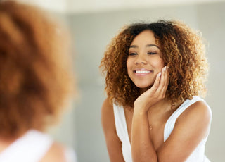 Female of color in a white tank top touching her cheek while examining her skin in the mirror. 