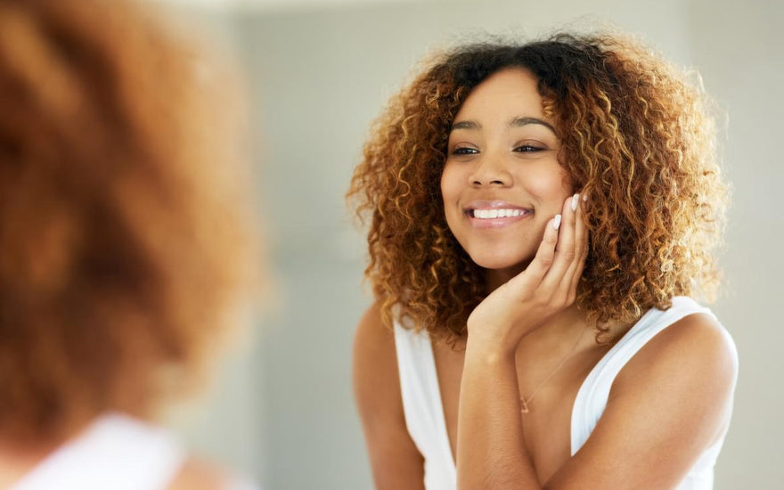 Female of color in a white tank top touching her cheek while examining her skin in the mirror. 