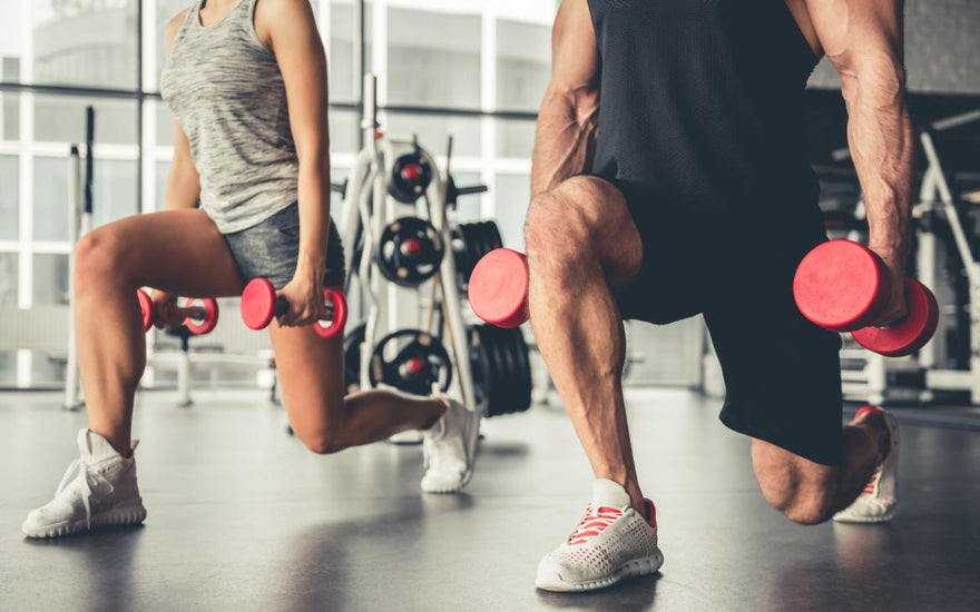 Couple doing lunges while holding red dumbbells in a gym. 