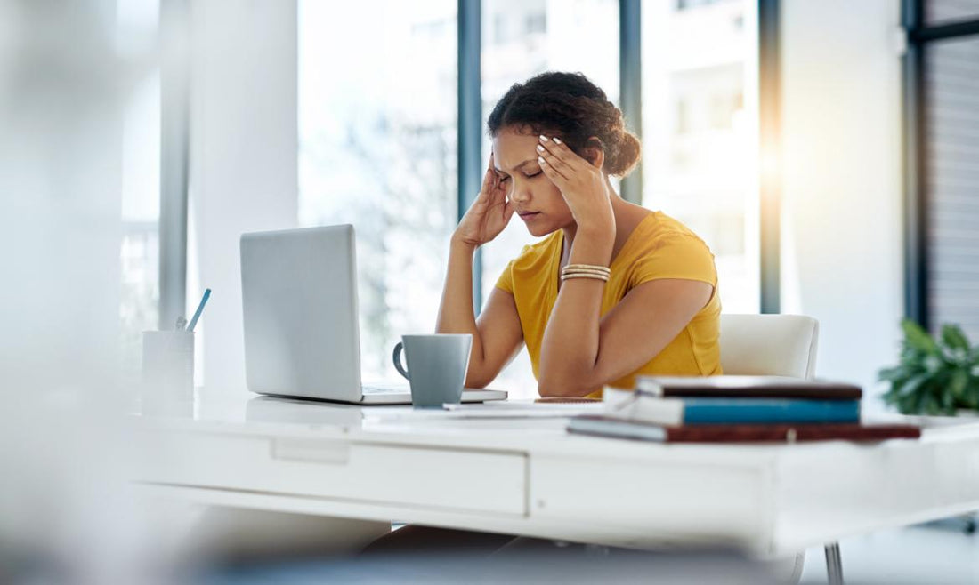 Women in a yellow blouse sitting at her desk with her laptop open. She has her fingers on her temples and her eyes closed.