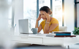 Women in a yellow blouse sitting at her desk with her laptop open. She has her fingers on her temples and her eyes closed.