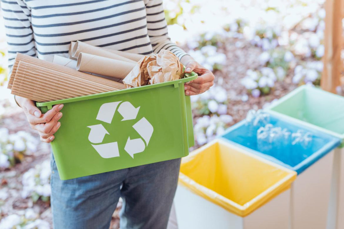 Woman holding a green recycling pin full of cardboard is standing frot of three color-coded waste bins.